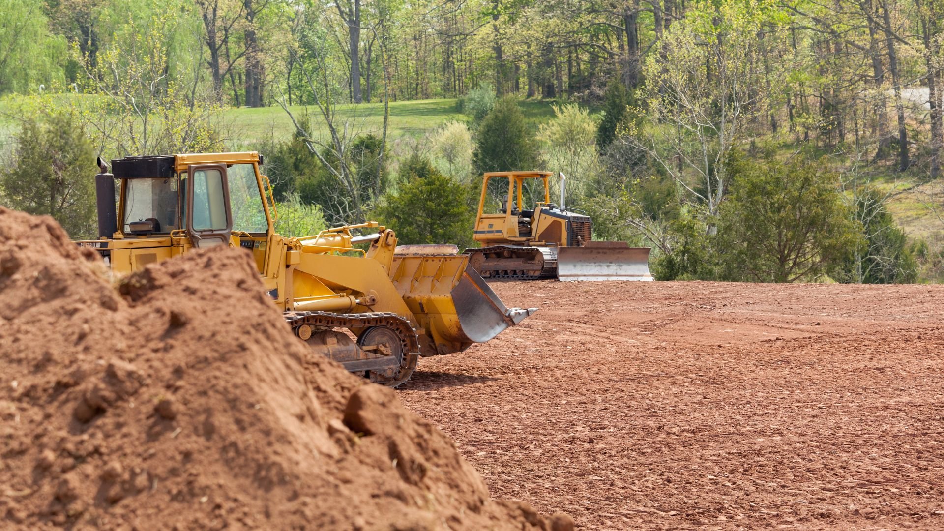 Yellow bulldozers clearing land in forested area with dirt piles